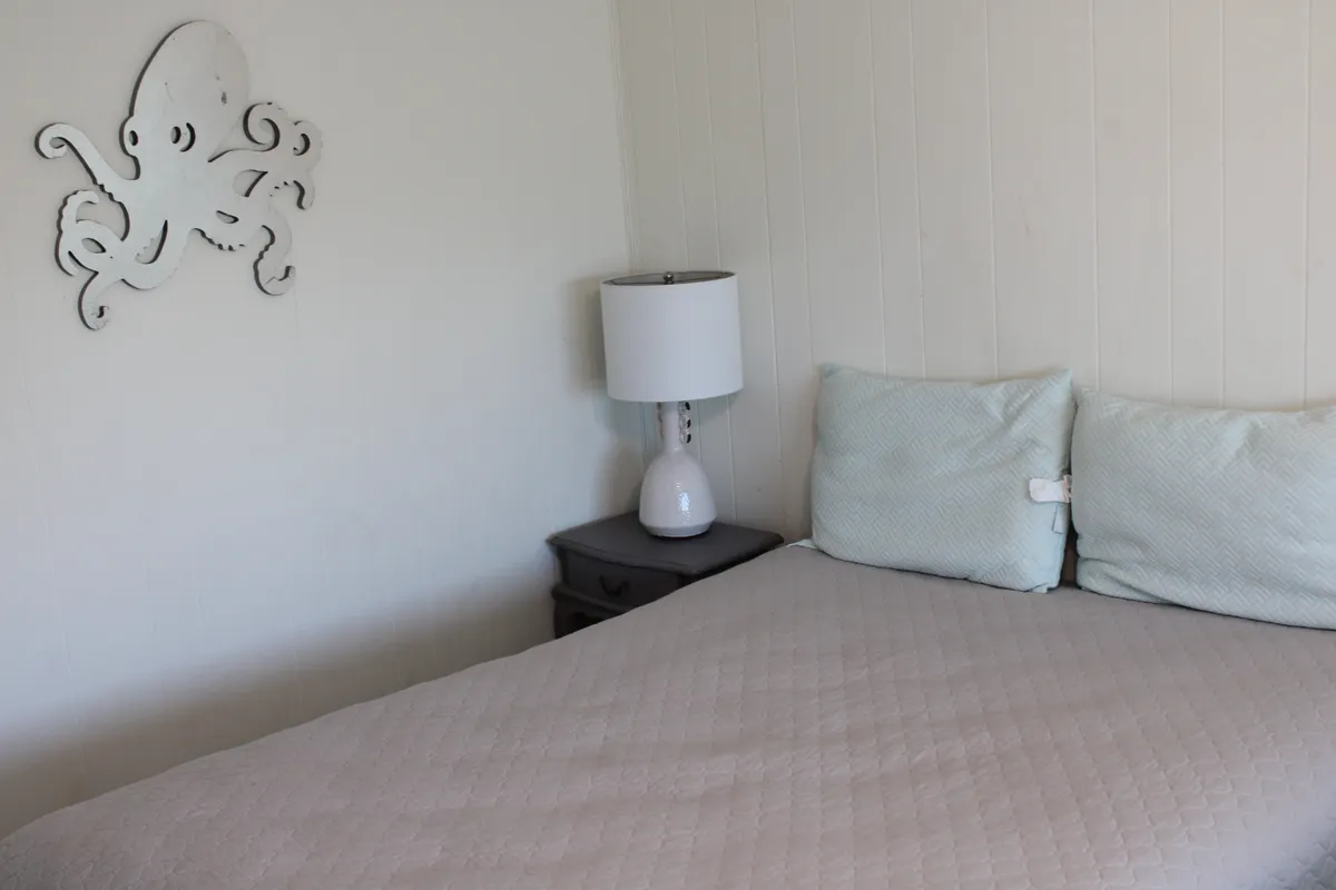 This image features a tranquil bedroom with light blue pillows on the bed, an inviting lamp, and octopus wall art on the wall. The decor is serene and ocean-inspired, evoking a peaceful, coastal retreat feeling perfect for couples seeking relaxation.