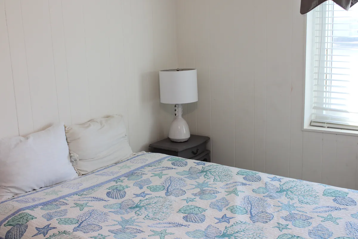 A cozy bedroom with a seashell-patterned quilt, a lamp on the nightstand, and a window with blinds that lets in gentle natural light. The scene suggests quiet mornings or comforting evenings, ideal for couples.