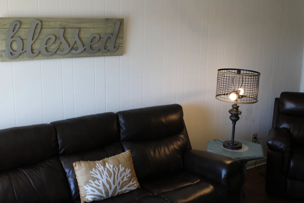 A comfortable living area with a leather sofa, decorative pillow, table lamp, and a wooden sign. The space is warm and inviting, perfect for relaxing together after a day of beachside exploration.