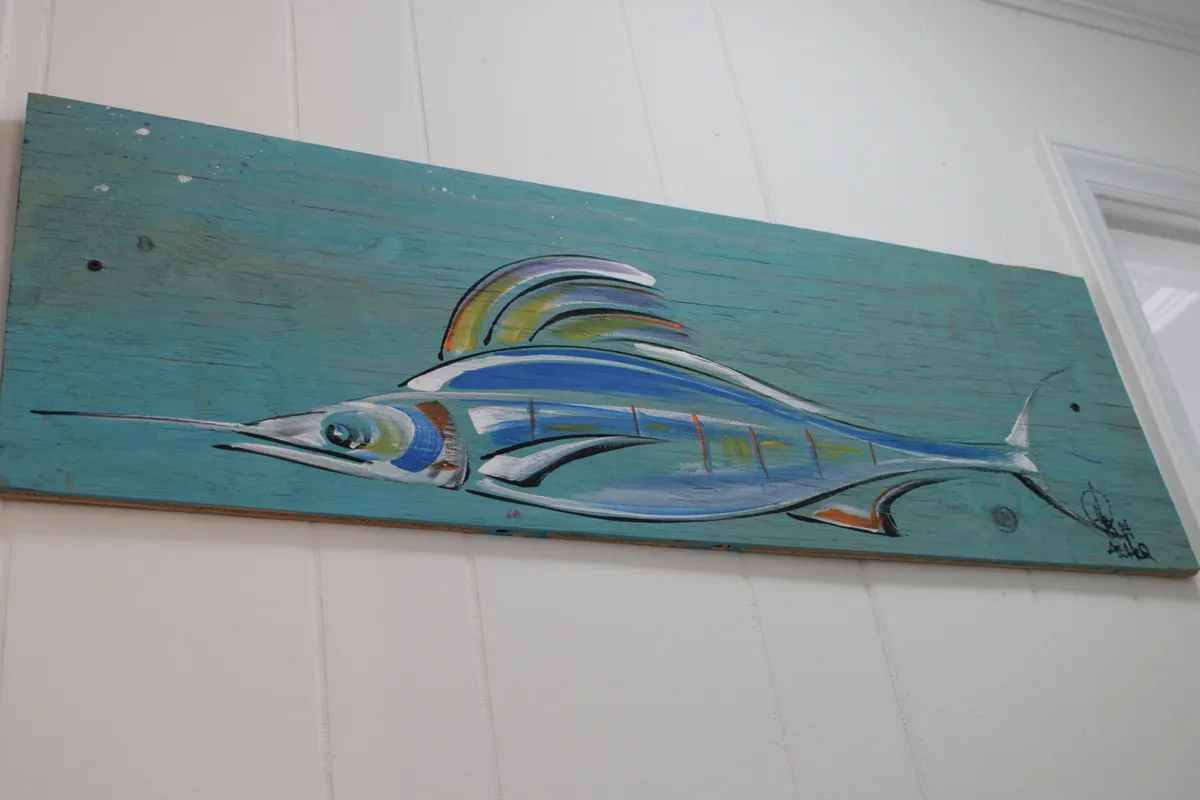 A cheerful coastal-themed decor scene featuring hand-painted fish artwork on a weathered wooden board. The bright, welcoming motif captures the local flavor and celebrates the beachside setting.