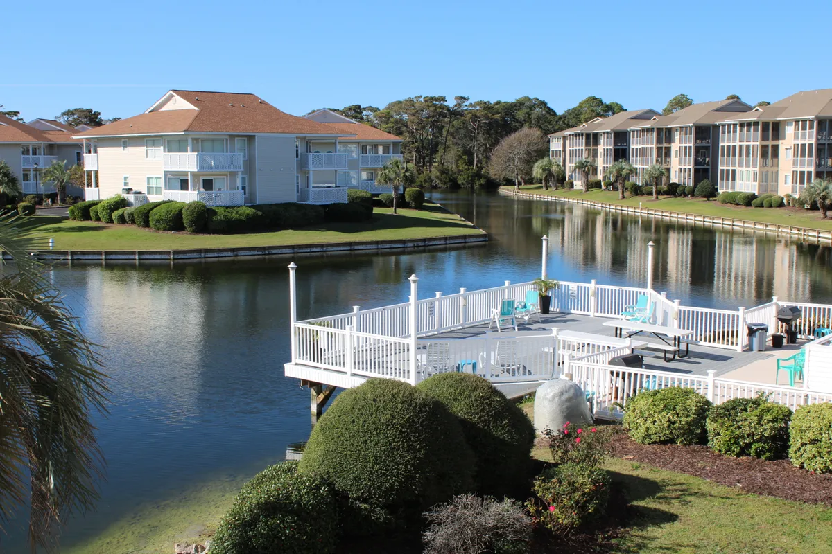 A calm dock with seating set against a serene body of water, surrounded by landscaped greenery and residential buildings featuring balconies. This outdoor scene evokes relaxation and a sense of community, with the balconies subtly connecting the private and communal aspects of the environment.
