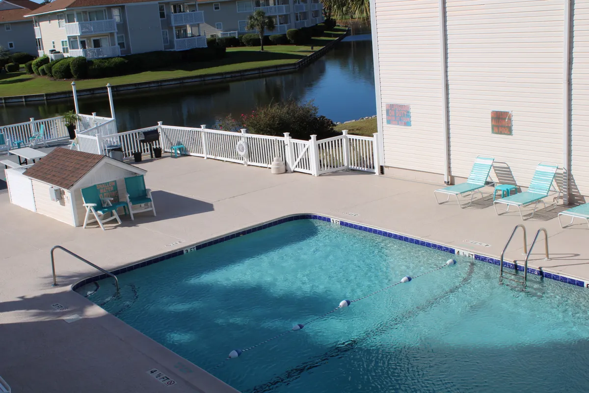 The image showcases a sparkling swimming pool with lounge chairs and a view of a nearby lake, all bordered by well-kept landscaping. The pool area is inviting for sunbathing and socializing while providing a clear sense of the community's tranquil heart.