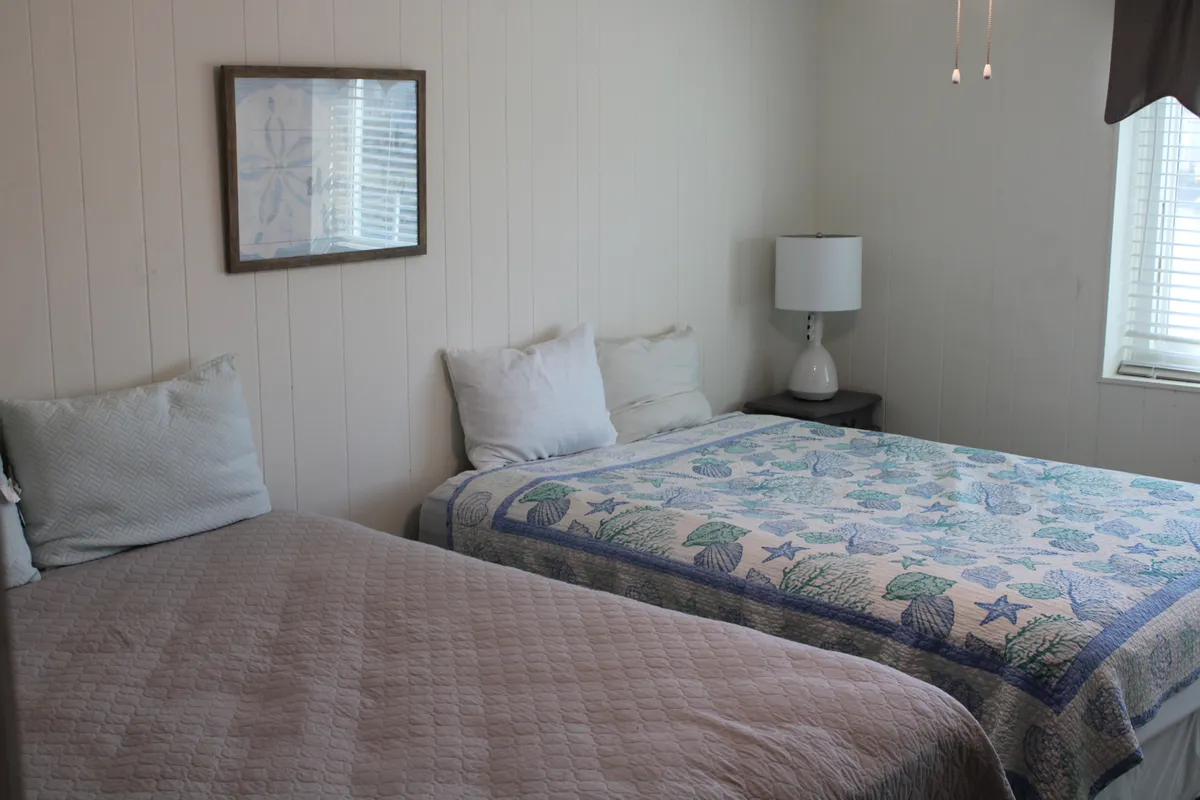 A cozy bedroom scene featuring two beds with decorative quilts, framed artwork, and a bedside lamp. The overall mood is relaxed and inviting, suggesting spaces for both family downtime and intimate escapes.