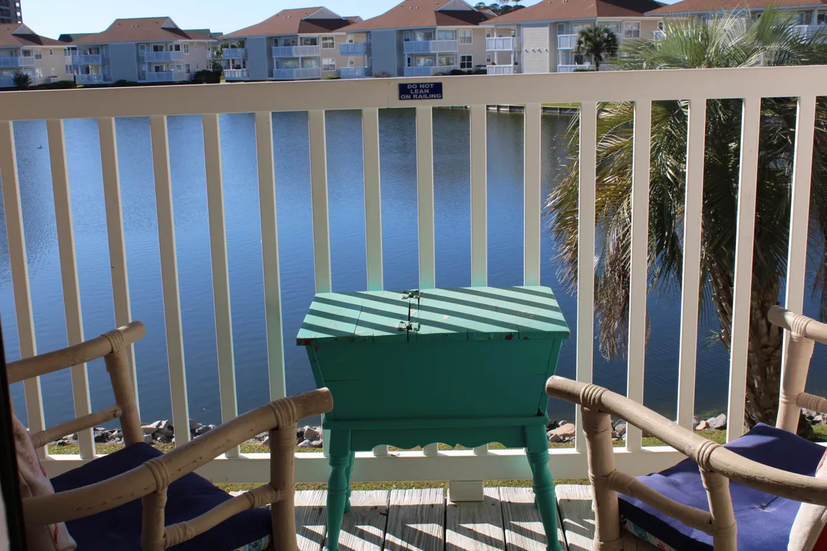This image features an inviting outdoor balcony space with a green table and wooden chairs, overlooking a tranquil lake framed by palm trees. The setting is peaceful, with the table arranged for relaxing or socializing while enjoying views of the water and lush surroundings.