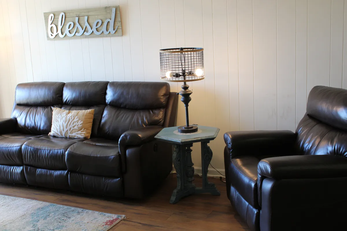 This image features a cozy living area with leather sofas, a decorative pillow, a table lamp, and wall art. The space is arranged to encourage conversation and relaxation, with natural light illuminating the room, creating a welcoming ambiance.
