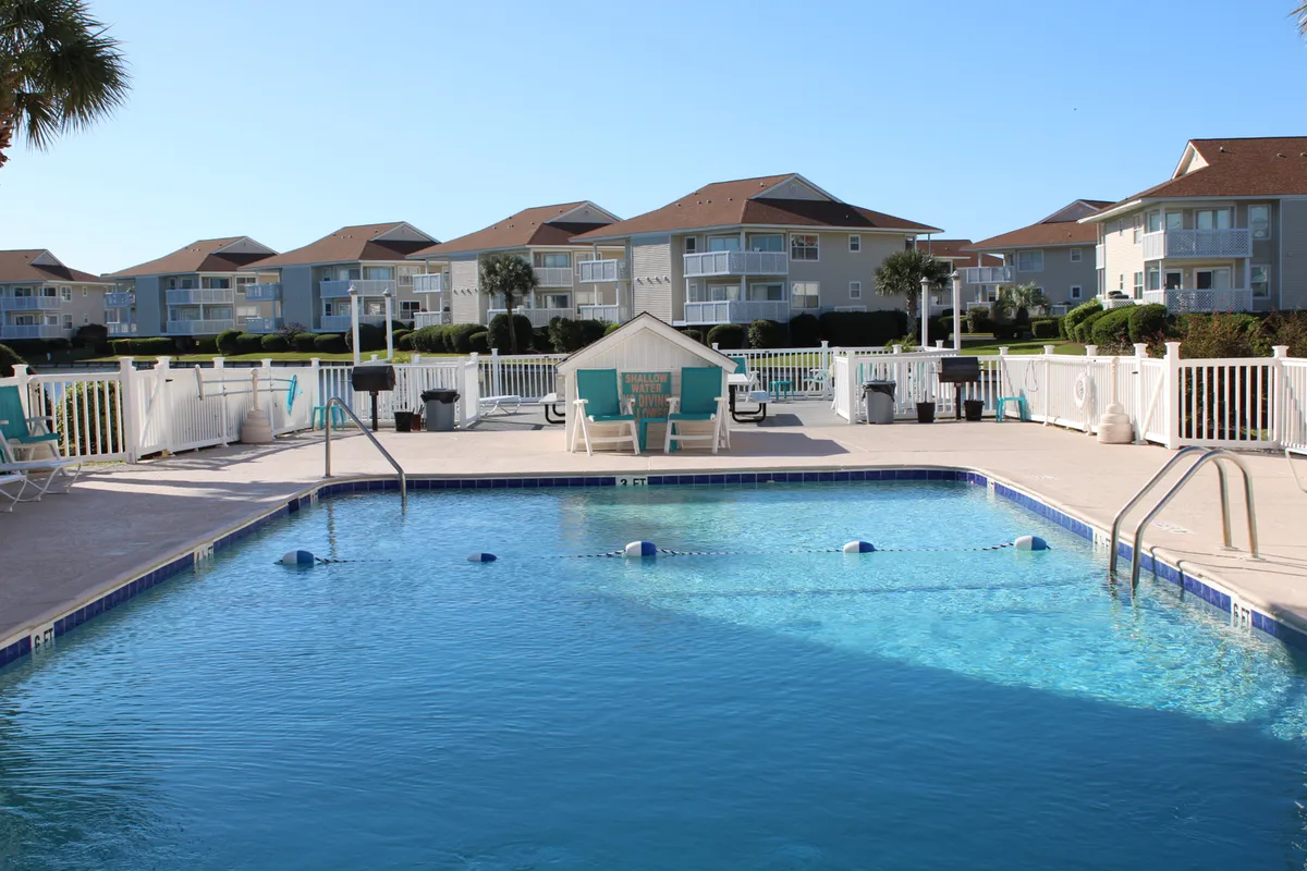This exterior scene features a large swimming pool surrounded by lounge chairs, a green lawn, and nearby buildings within the Beach Cottages community. The area includes a barbecue area and is designed for both relaxation and social gatherings. The overall mood is inviting and serene, perfect for families returning from the beach or unwinding under the sun.