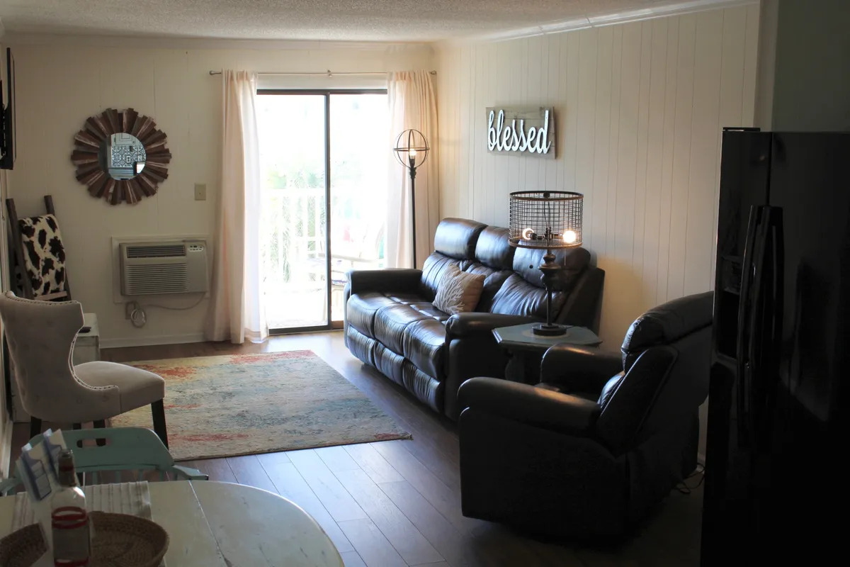 This cozy living area features leather sofas arranged for conversation, a decorative mirror, a 'Blessed' sign, and a side table with a warm lamp. Soft lighting and thoughtful décor elements create an inviting atmosphere perfect for relaxation after a day at the beach.